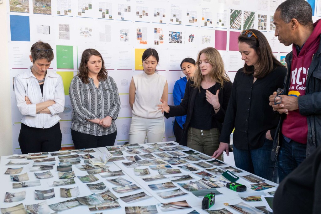 7 people stand around a square table covered with dozens of small color photographs.