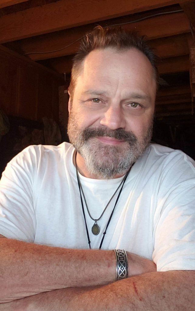 Headshot of Daniel Sayers who wears a white t-shirt and two necklaces.