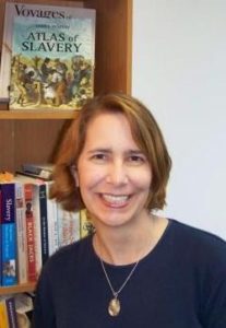 A white woman with short blond hair wearing a navy crewneck sweater and smiling, stands in front of a bookshelf