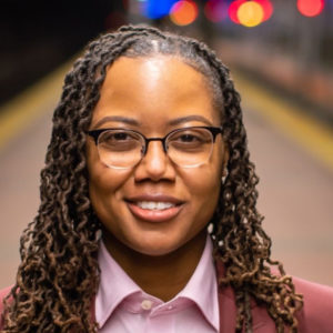 A Black woman wearing a pink shirt, darker pink jacket, and glasses, with long twisting braids, looks into the camera. Out of focus behind her is a train platform.