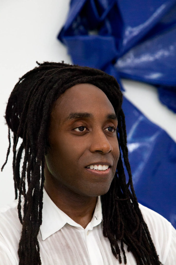 Headshot of Anthony Titus, who wears a white shirt and stands in front of a blue sculpture on a white wall.
