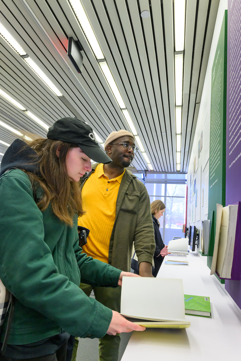 Two people looking at the long shelf of books on display in front of an exhibition wall of book covers and abstracts
