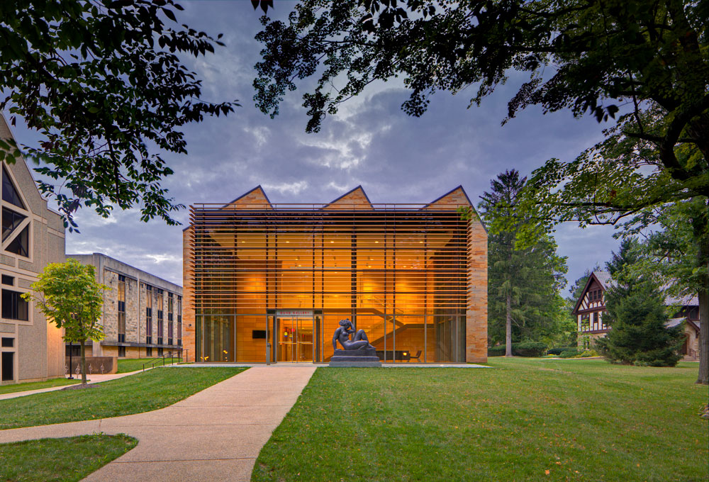 Illuminated gallery building viewed from the exterior, with grass, trees, and a statue.